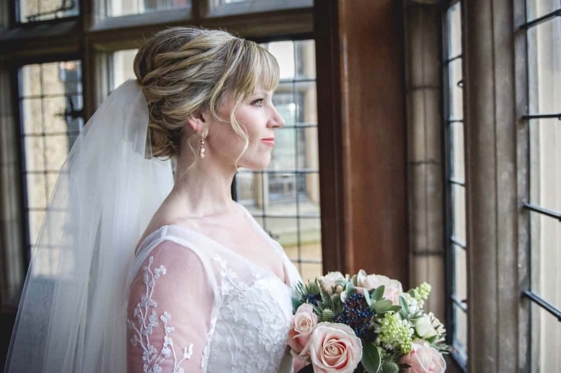 Bride with braided updo looks out of window of Fanhams Hall, Ware, holding bouquet